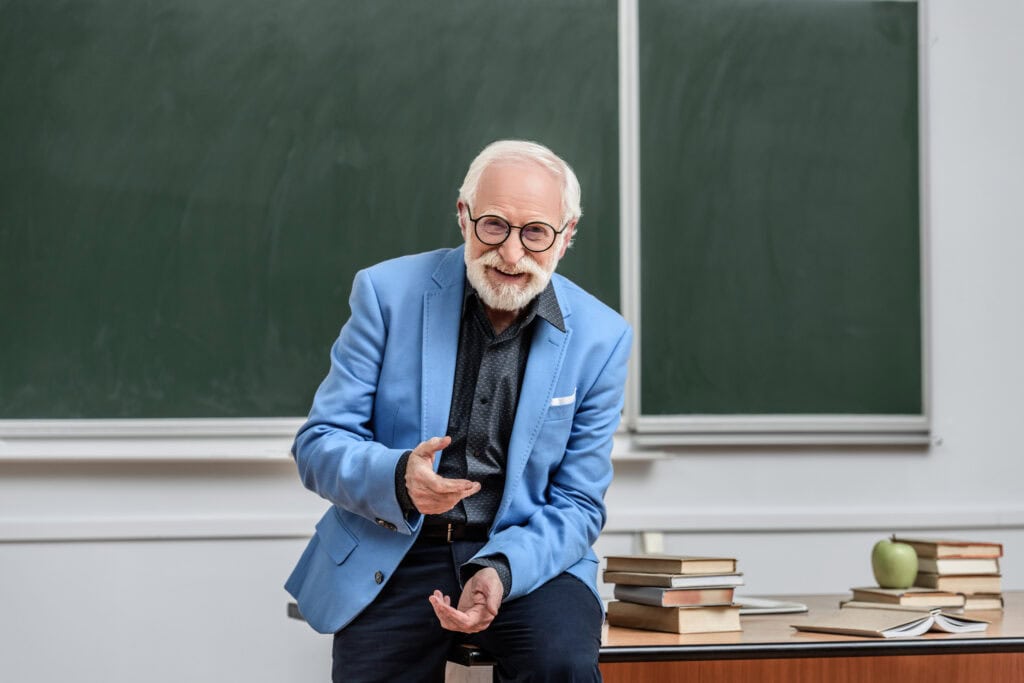 smiling grey hair professor sitting on table in lecture room