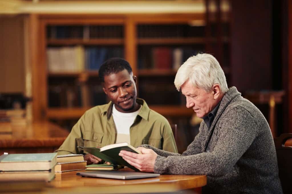Portrait of male senior student reading book in library while preparing for exam with a friend